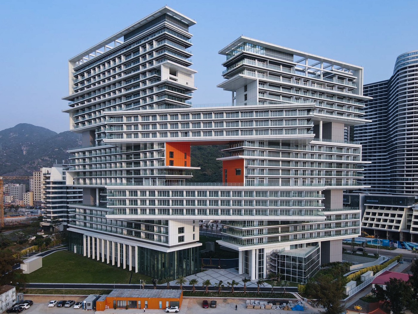 Modern, multi-level glass building with stacked terraces and orange accent panels, set against a cityscape and hills in the background.