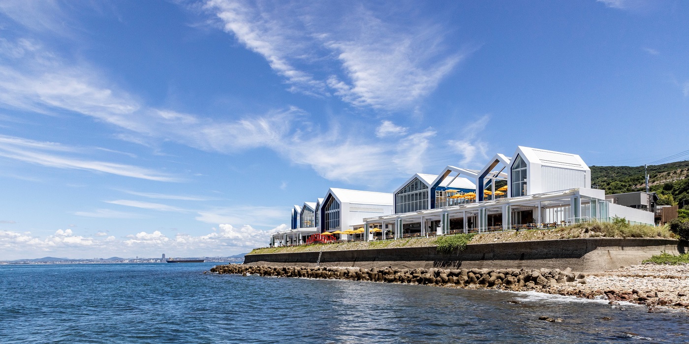 Row of white gabled buildings along a rocky coastal seawall, overlooking calm blue water under a clear sky with wispy clouds.