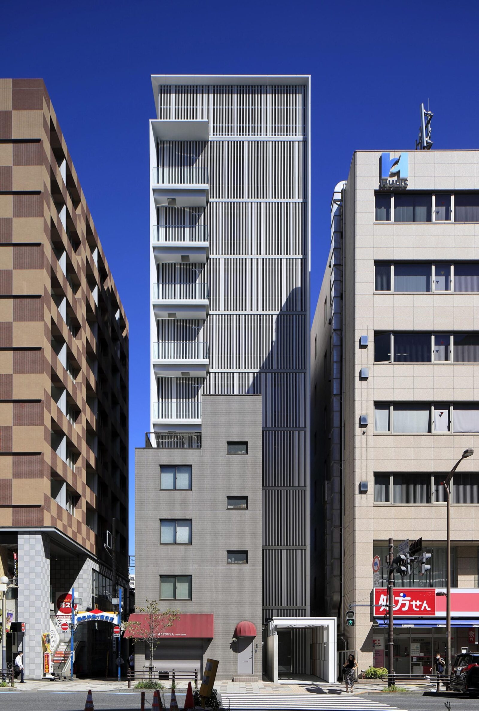 Tall modern white high-rise with vertical louvers between brown brick building on the left and beige office block on the right, under a clear blue sky.