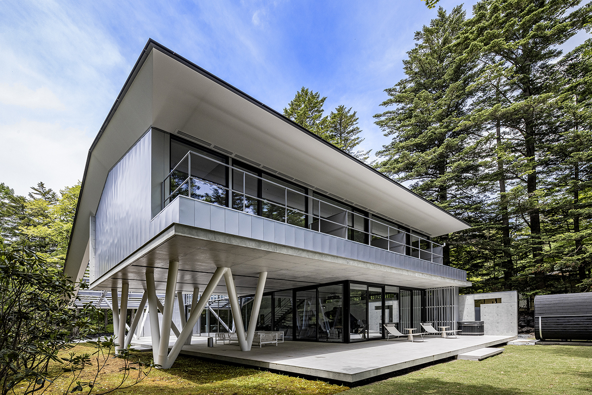 Contemporary two-story house with a cantilevered upper level, glass walls, and angled white support beams on a sunny lawn surrounded by trees.