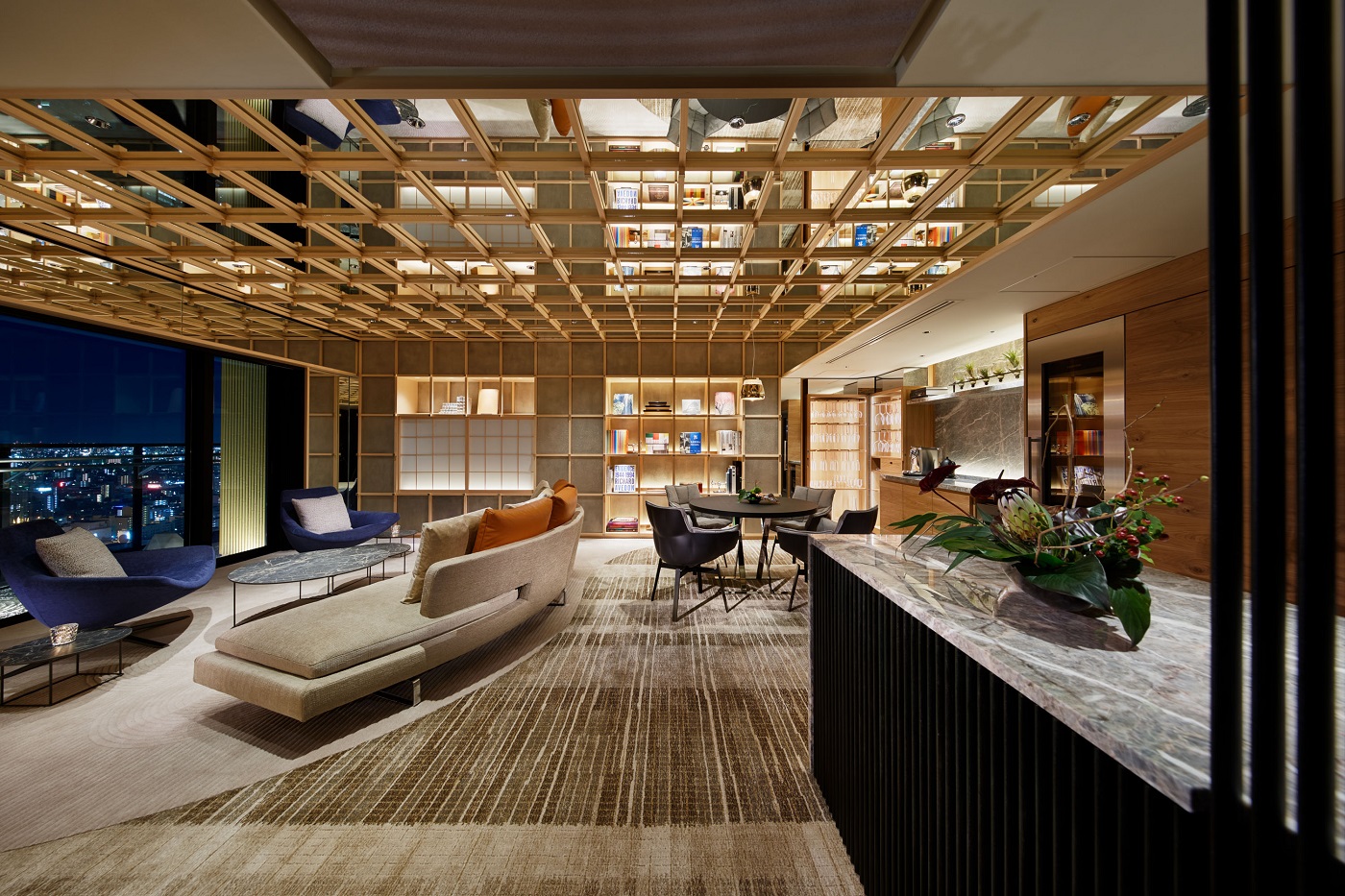Modern hotel lounge with beige sectional sofa, blue lounge chairs, and round marble coffee tables, facing a wall of built-in bookshelves.