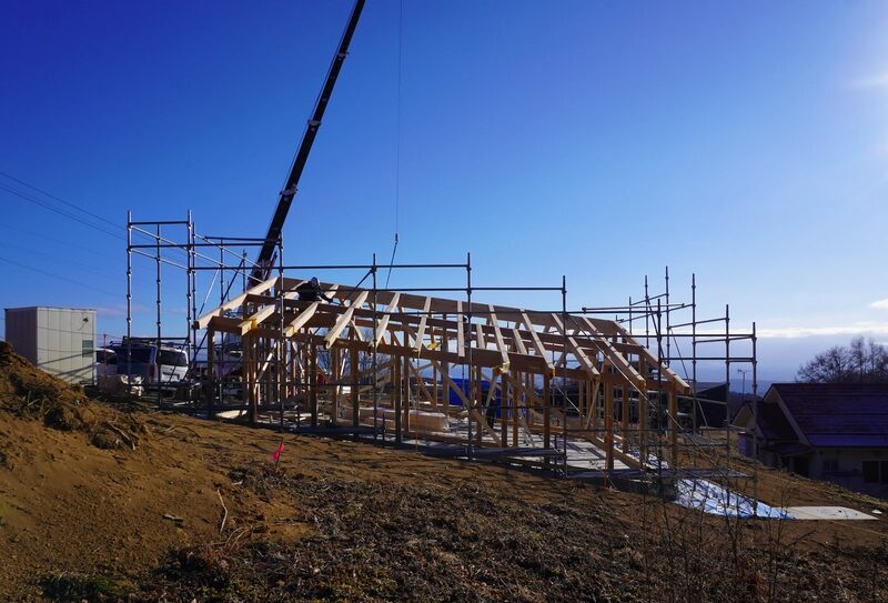 Wood-framed building under construction with scaffolding on a dirt site and a crane overhead.