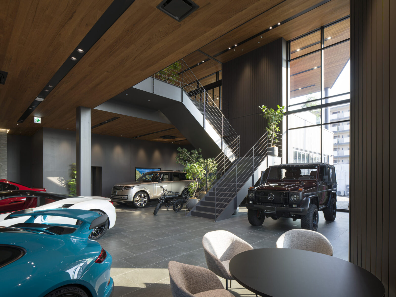Interior of a modern car showroom with a wooden ceiling, metal staircase, and several luxury cars on display by a large glass wall.