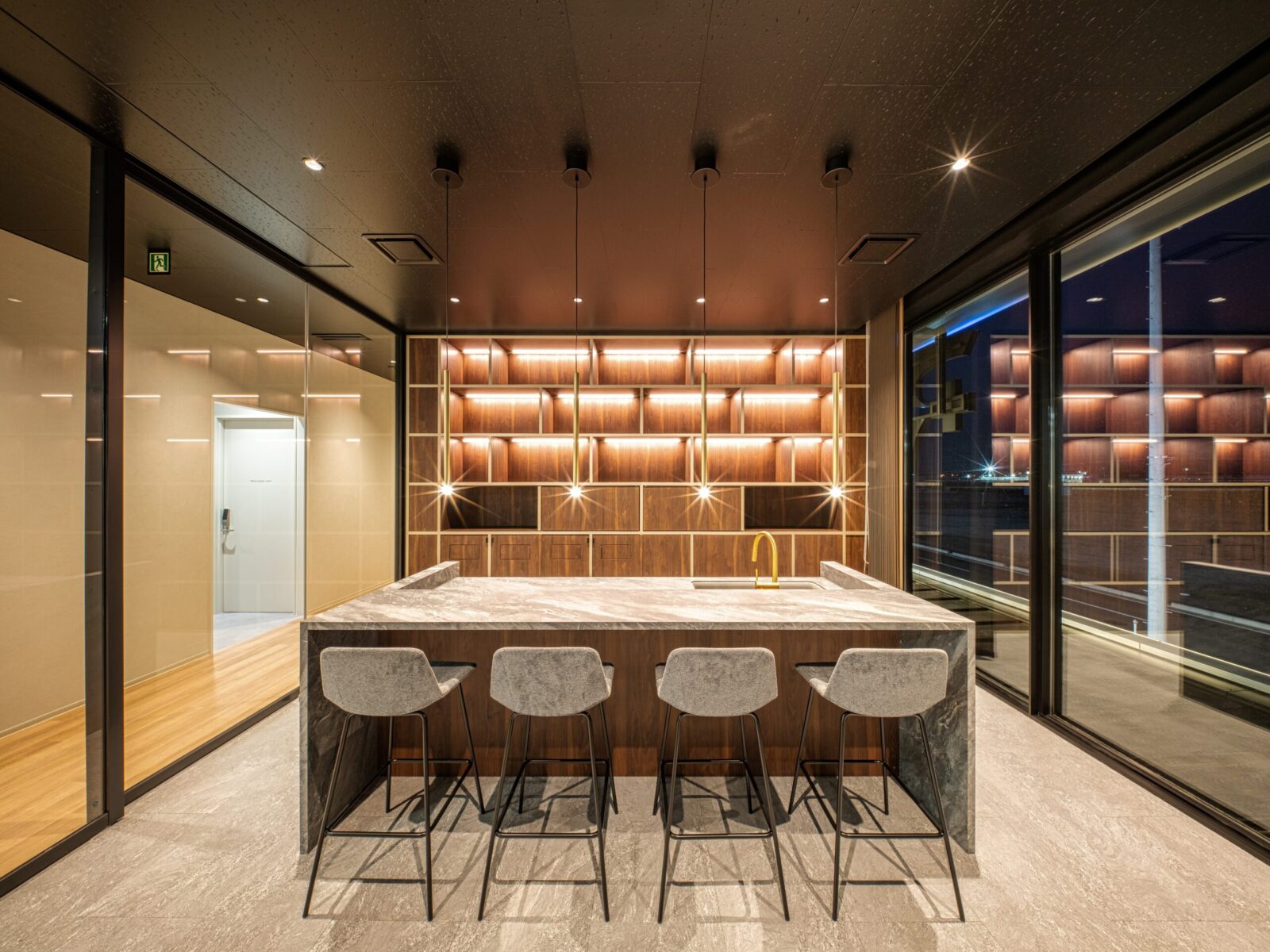 Modern kitchen with a marble island, four gray bar stools, and backlit wooden shelves in warm lighting at night.