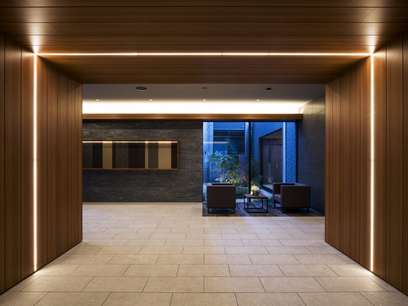 Wood-clad hallway opening into a lobby with a seating area beyond a glass wall and blue exterior light.