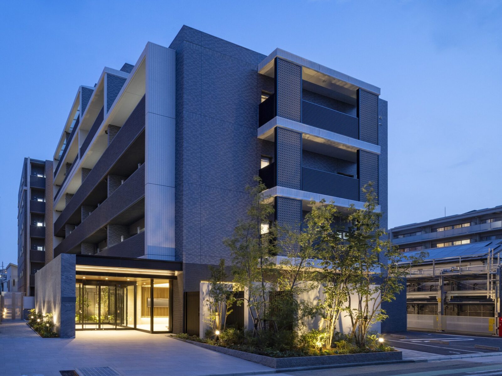 Modern multi-story apartment building at dusk, with lit ground-floor lobby and landscaped entrance.