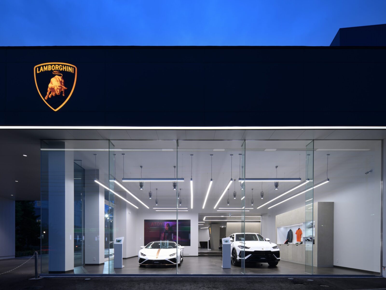 Lamborghini showroom exterior with a lit orange emblem on a dark wall and glass storefront revealing white sports cars inside