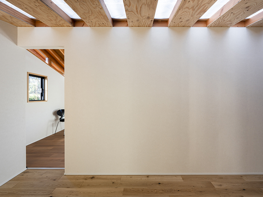 Minimalist interior with white walls, wooden floor, and exposed timber ceiling beams; doorway reveals a sunlit room with a window and chair.