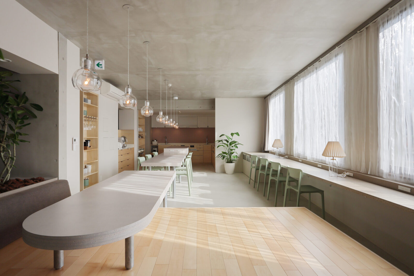 Bright cafe interior with long white tables, mint-green chairs, and glass pendant lights along the center.