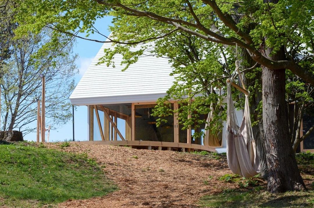 Modern glass-walled house on a grassy hillside, with a wooden deck and a hammock tied to a tree.
