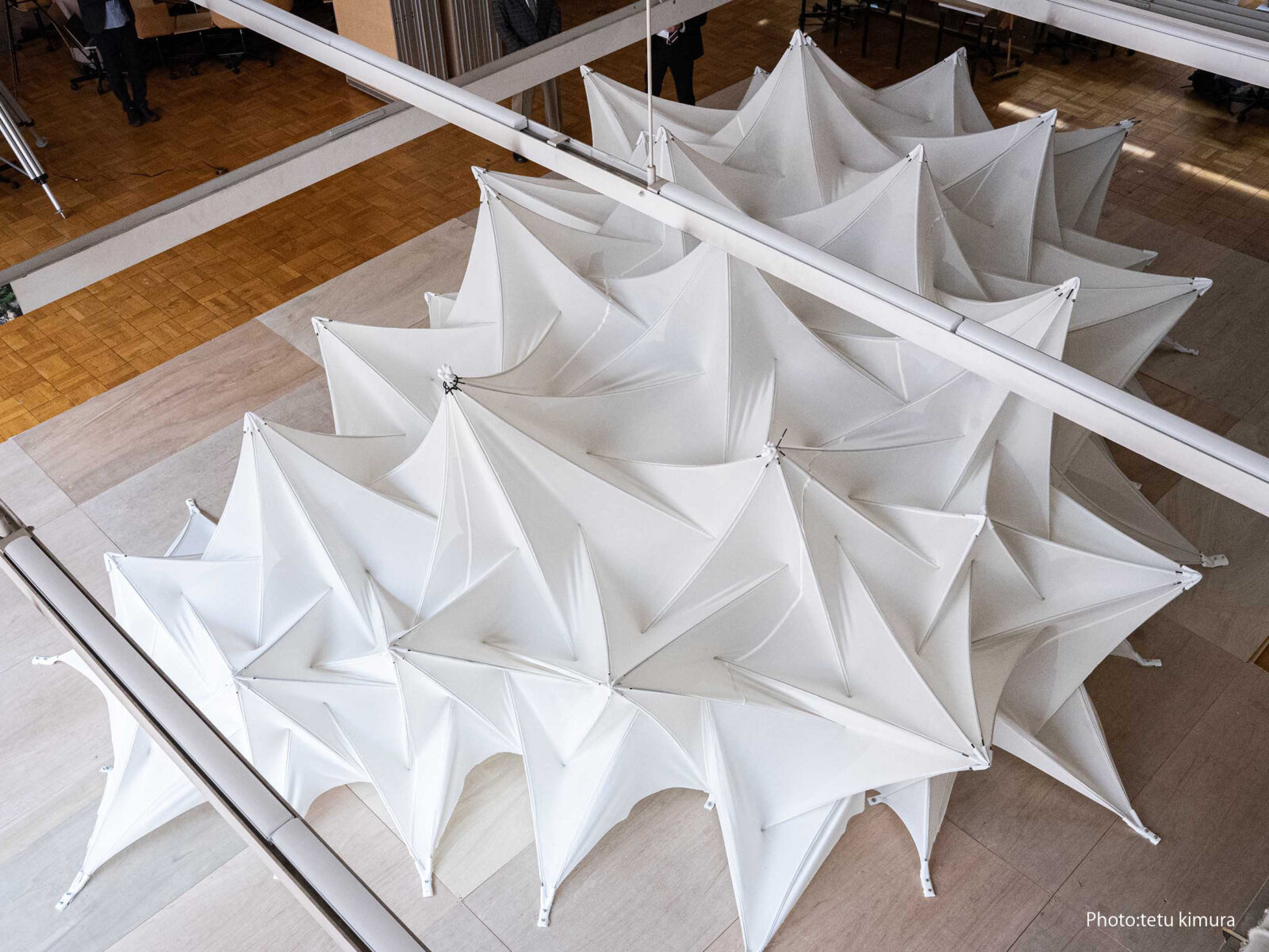 Indoor art installation of white geometric canopies laid out on a wooden floor, viewed from above.