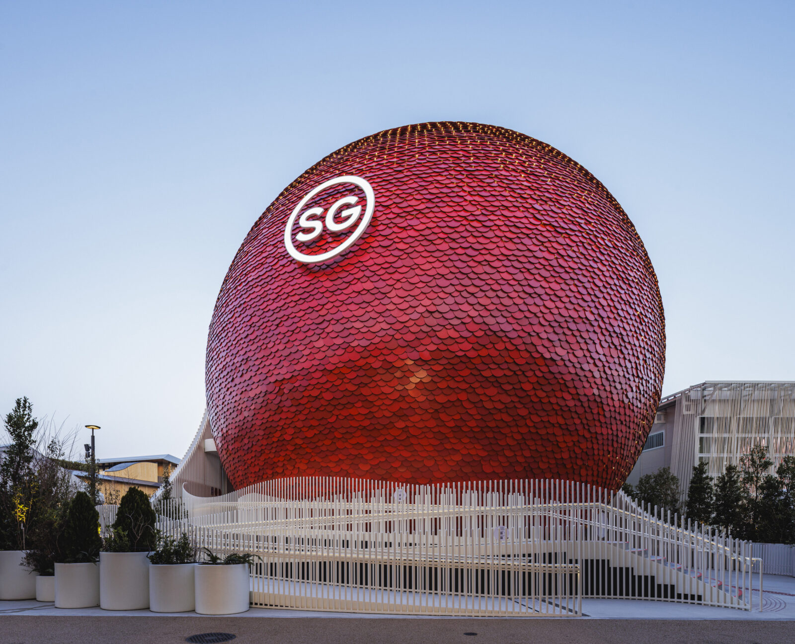 Large red spherical building clad in small scale tiles, with a white circular SG logo on its surface, surrounded by a white fence and trees against a blue sky.