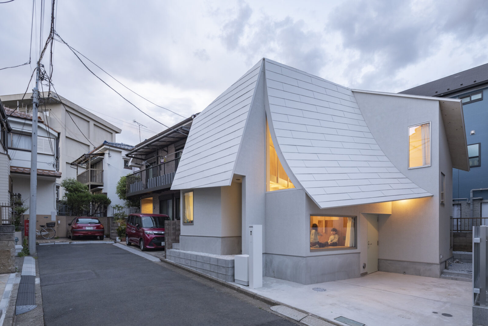 Modern house with a dramatic triangular, silver roof and warm-lit windows on a residential street.