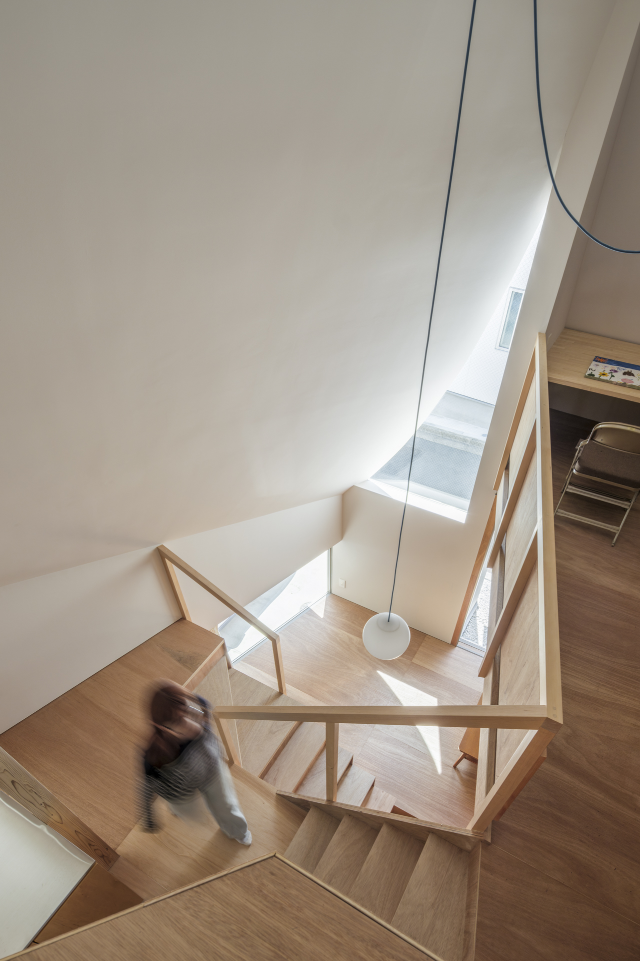 Interior view of a modern wooden staircase in a bright atrium, with curved white walls and a hanging pendant light, a blurred person on the stairs."
