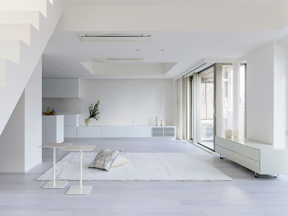 Minimalist white living space with a white rug, light furniture, and large floor-to-ceiling windows on the right.