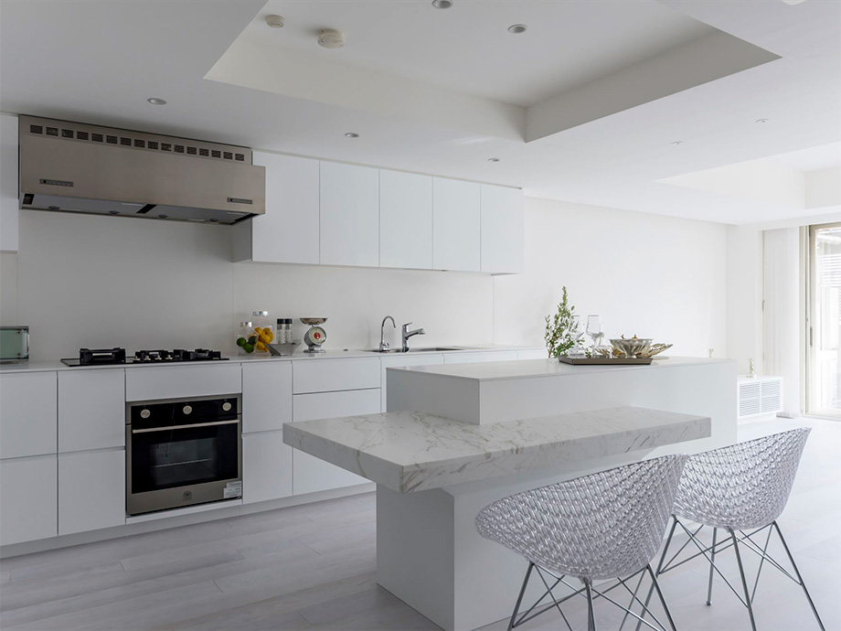 Bright white modern kitchen with a marble island, gas cooktop, built‑in oven, and white cabinetry, plus a stainless range hood on the left.