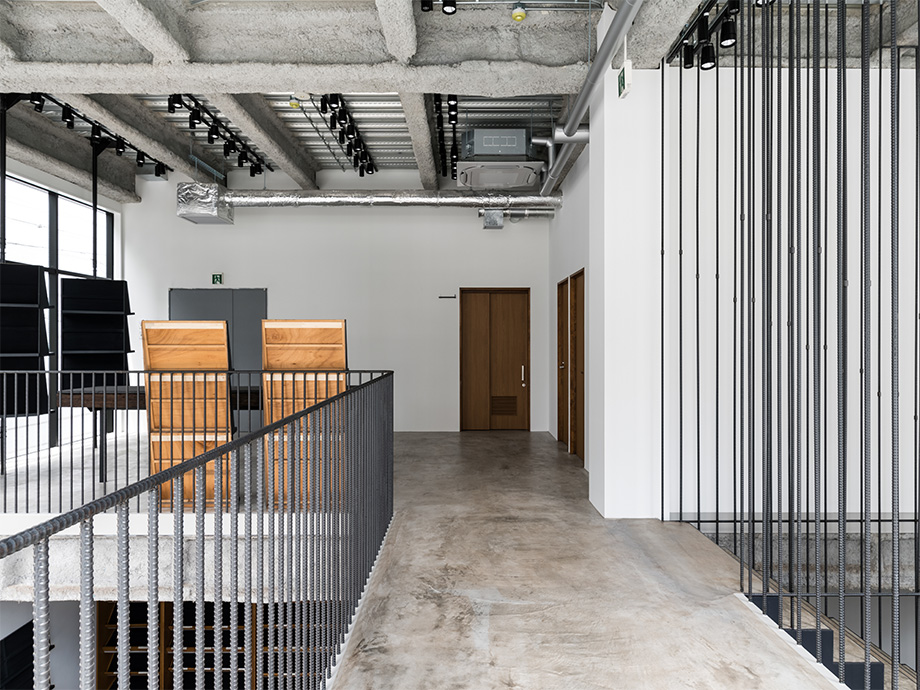 Industrial loft gallery space with exposed concrete ceiling, track lighting, and a metal railing along a walkway; wooden display boards on the left and vertical metal bars along the right, with doors at the far end.