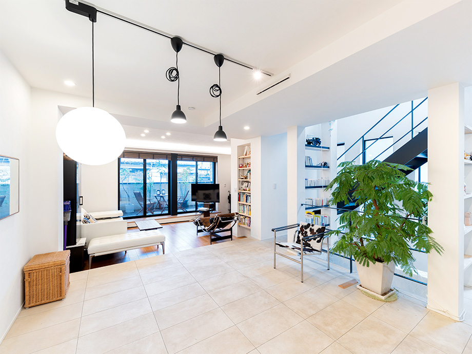 Bright open living area with white furniture, a large potted plant, and a glass door opening to a patio. Two-tier bookshelves and a staircase are visible in the background.