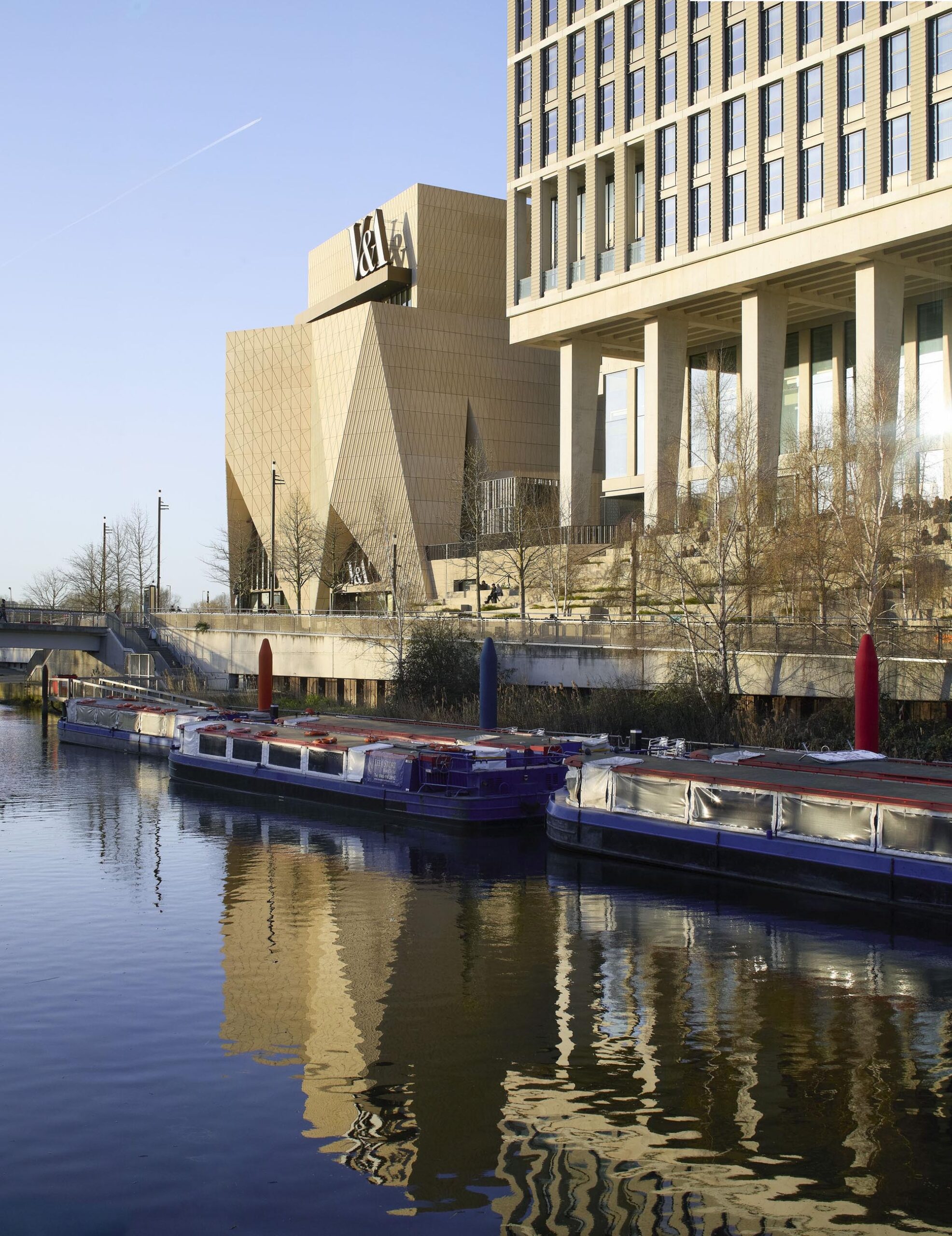Modern beige brick and glass building along a calm river with reflected façades and moored boats in the foreground.