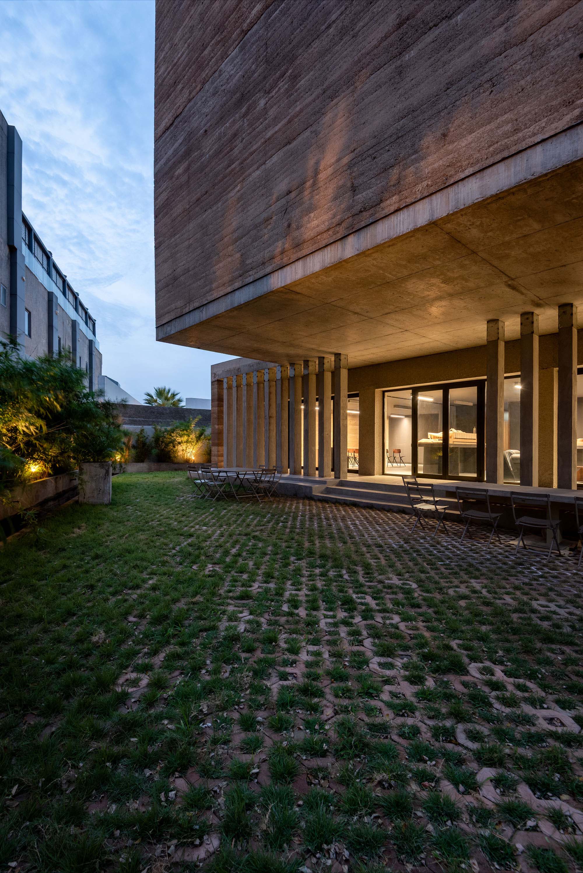 Modern concrete building with a large cantilevered upper level and glass entrance; outdoor seating under exposed columns at dusk.