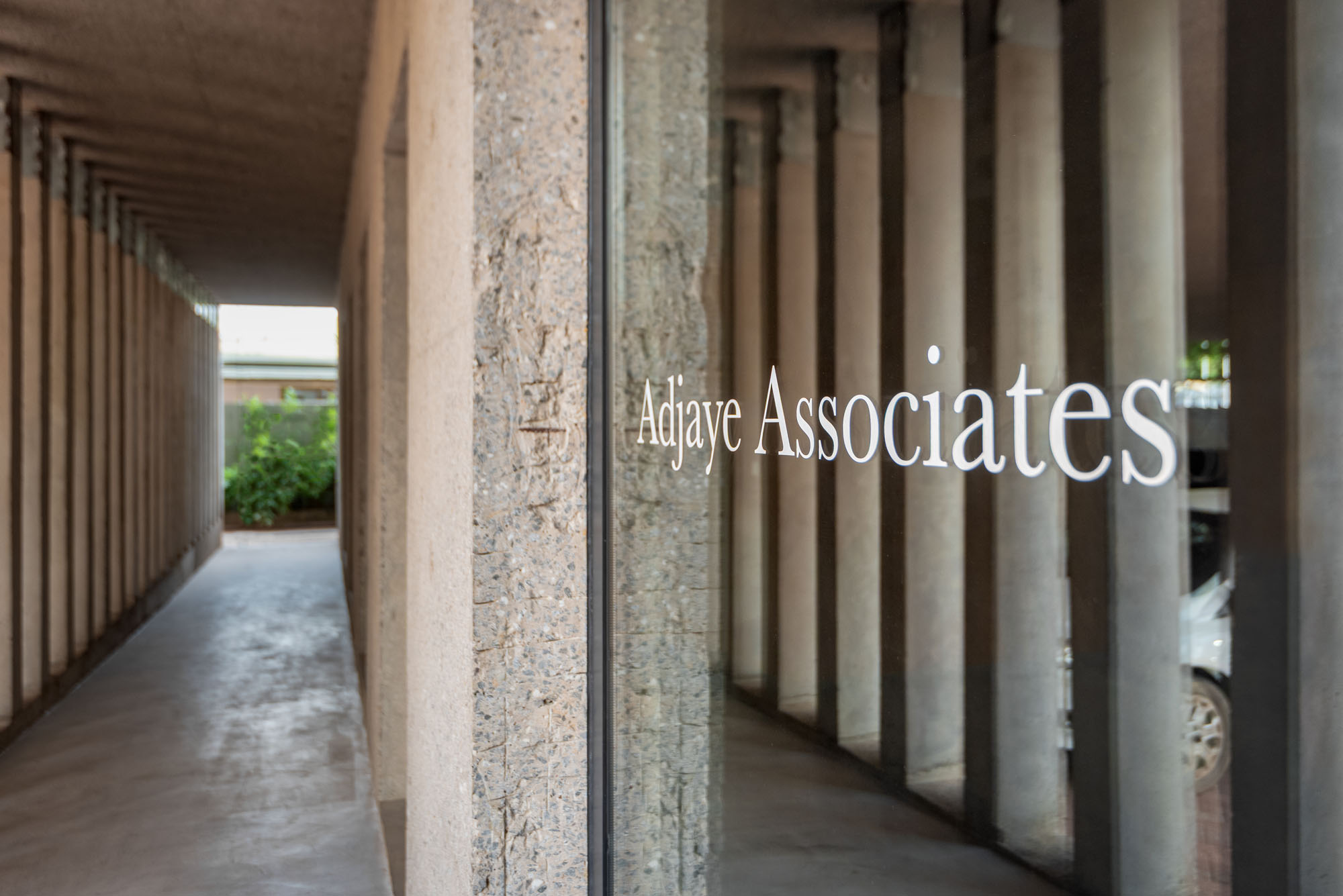 Clear glass window showing the name 'Adaye Associates' along a stone wall, with an alleyway of columns visible beside it.