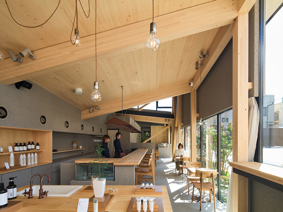 Bright cafe interior with wooden beams, a long communal counter, bar stools, and large windows showing sunny seating areas.