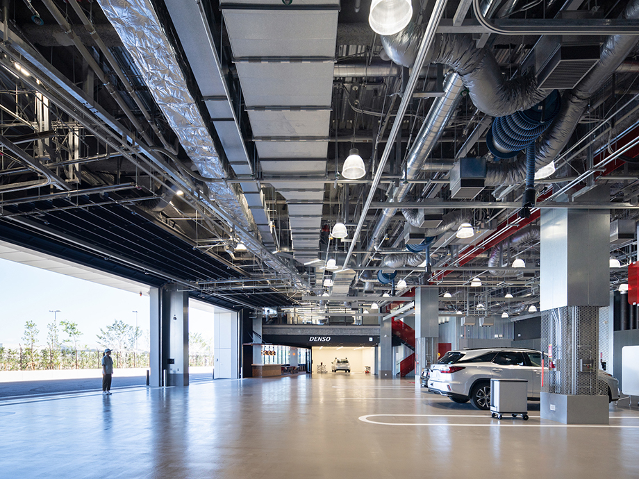 Open, spacious automotive service bay with exposed pipes and ducts overhead; a person stands near the left entrance while a white SUV is parked inside.