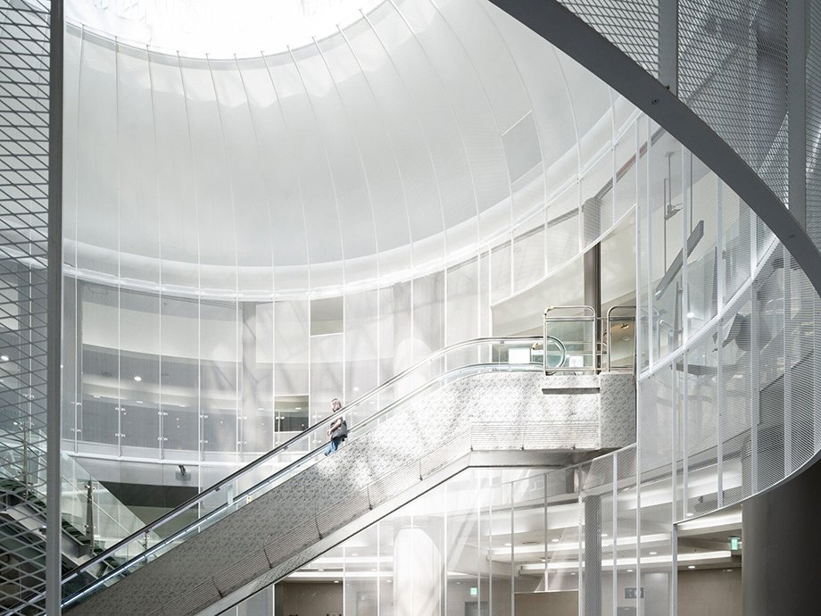 Curved glass atrium with an escalator rising through a bright, modern interior building.