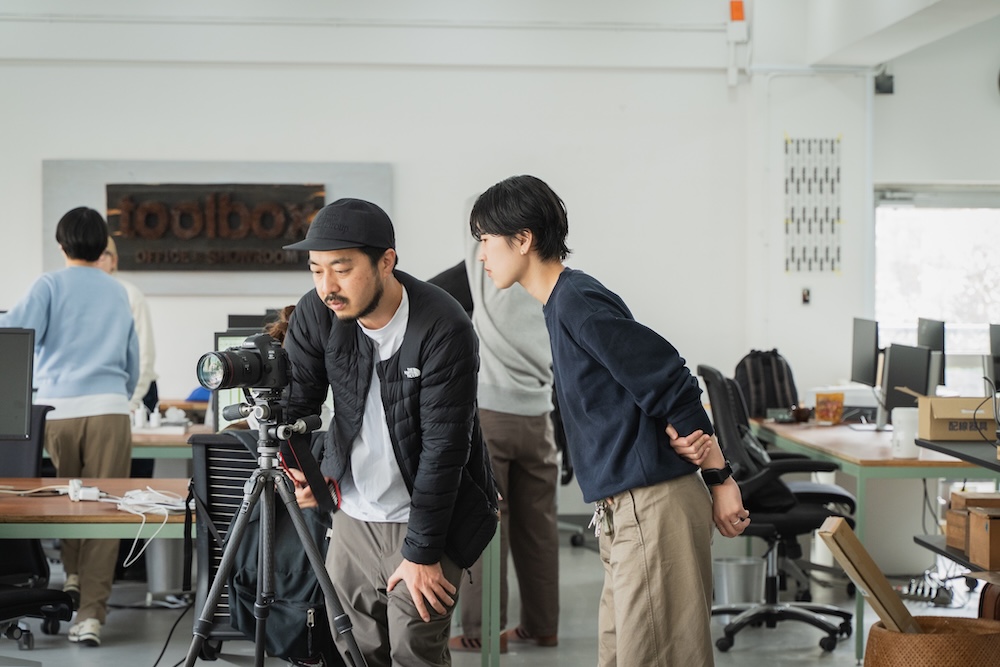 Two men in a bright office lean over a camera on a tripod, inspecting the setup together.
