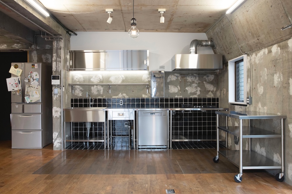 Industrial-style kitchen with stainless steel appliances, black tiled backsplash, concrete walls, and a mobile steel cart on wheels in the foreground.