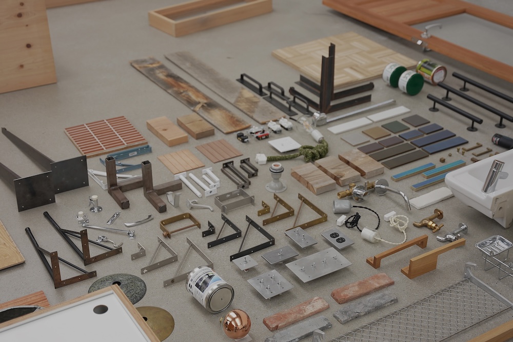 Wide assortment of hardware and building samples laid out on a workshop floor, including metal brackets, wood tiles, pipes, and small tools ready for display or selection.